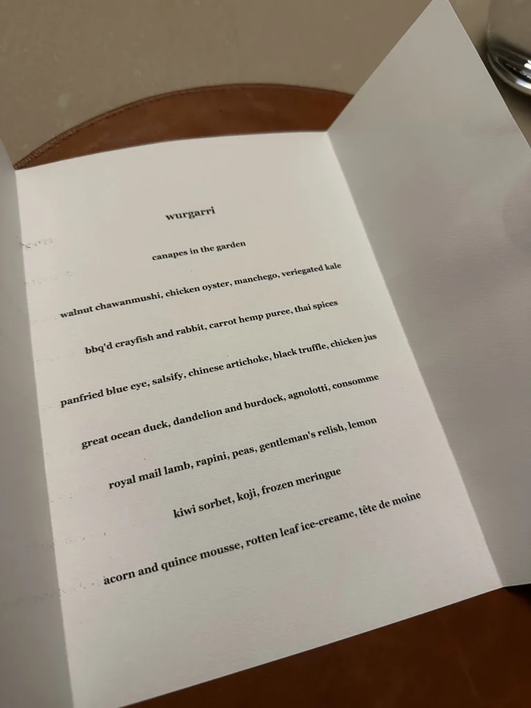This image shows a printed menu card titled "wurgarri," detailing a sequence of courses starting with "canapes in the garden". The list features sophisticated dishes such as bbq'd crayfish and rabbit, panfried blue eye, royal mail lamb, and acorn and quince mousse.