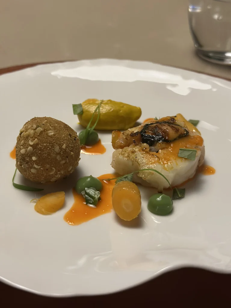 This vibrant dish showcases a golden-brown, seed-crusted croquette paired with a succulent piece of seafood topped with a rich orange glaze. The composition is artistically arranged with a smooth yellow quenelle, bright green peas, and splashes of vivid orange oil on a wavy white plate.