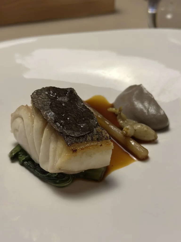 This refined dish features a flaky white fish fillet crowned with a slice of black truffle, resting on a bed of dark cooked greens. Adjacent to the fish lies a smooth quenelle of grey puree and tender stalks, all brought together by a rich, glossy amber sauce.