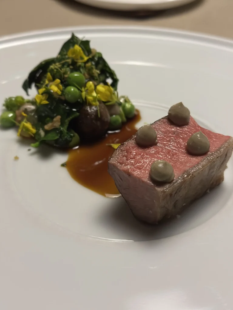 This savory dish showcases a tender, pink cut of meat garnished with four precise dollops of creamy puree. Accompanying the protein is a vibrant medley of greens, peas, and yellow flowers, resting in a pool of rich, glossy reduction sauce.