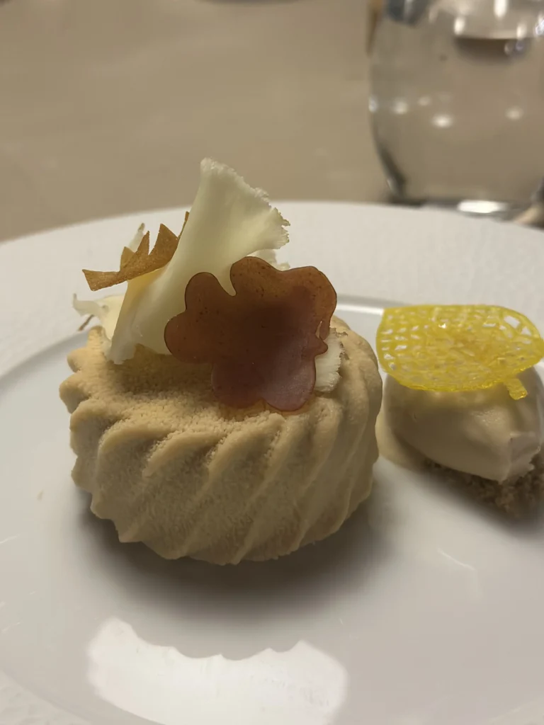 This gourmet dessert features a ridged, light brown mousse cake topped with white chocolate shards and a translucent amber crisp. Accompanied by a smooth quenelle of cream on a crumb base, the dish is accented with a delicate, bright yellow honeycomb tuile.