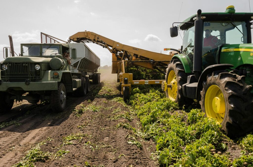 In a sunlit field, a large green tractor pulls a yellow harvesting implement alongside a rugged, olive-green truck.