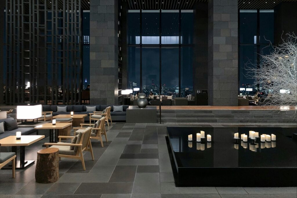 This image depicts a spacious, upscale lounge with high ceilings, dark stone columns, and large windows looking out onto a city at night.