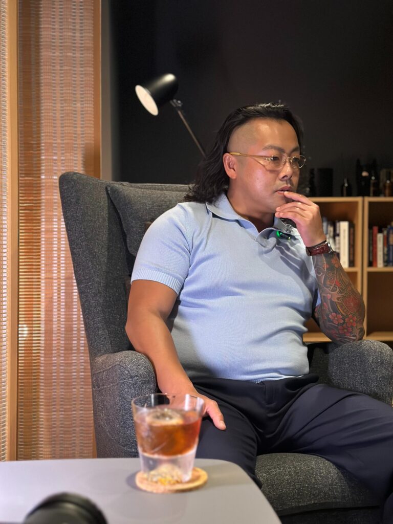 A man with a tattooed sleeve and glasses sits in a gray armchair, looking off to the side with a contemplative expression. In the foreground, a glass containing an amber liquid rests on a table, setting a relaxed, lounge-like atmosphere.