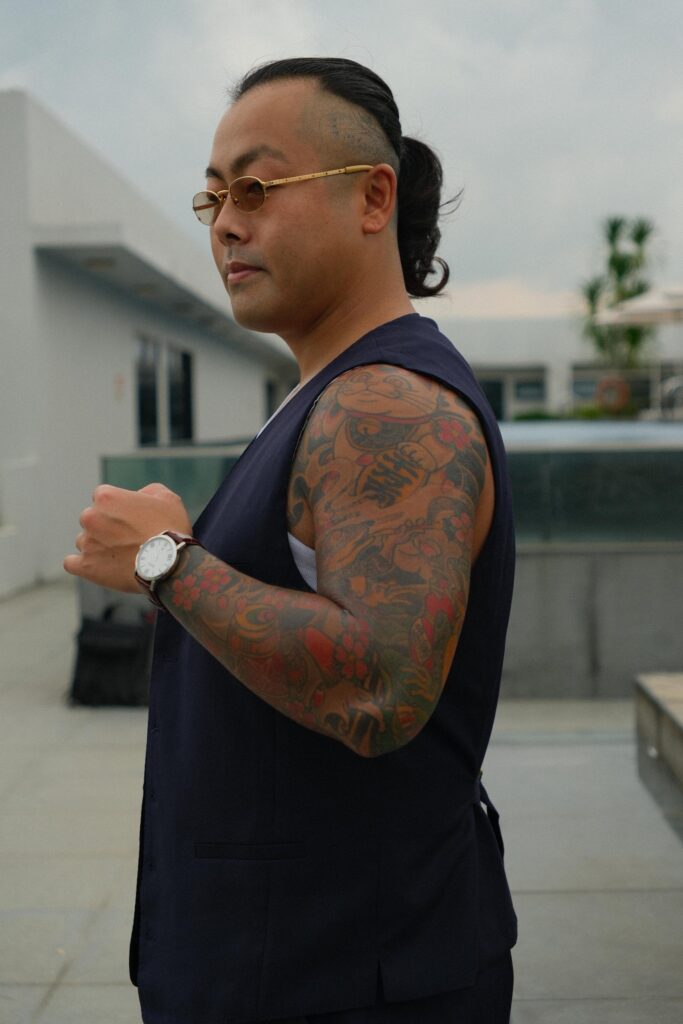 A man with a ponytail and gold-rimmed glasses stands in profile, wearing a dark blue vest over a white undershirt. His left arm is prominently displayed, showcasing a vibrant full-sleeve tattoo that features a Japanese lucky cat design.