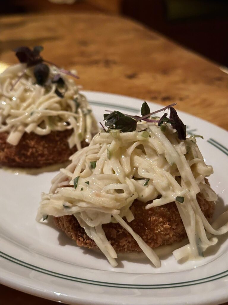 Two golden-fried cakes are topped with a generous mound of creamy shredded slaw and garnished with dark microgreens on a white plate with green trim. The close-up angle highlights the textures of the crispy breading and the fresh vegetable topping against a blurred wooden background.