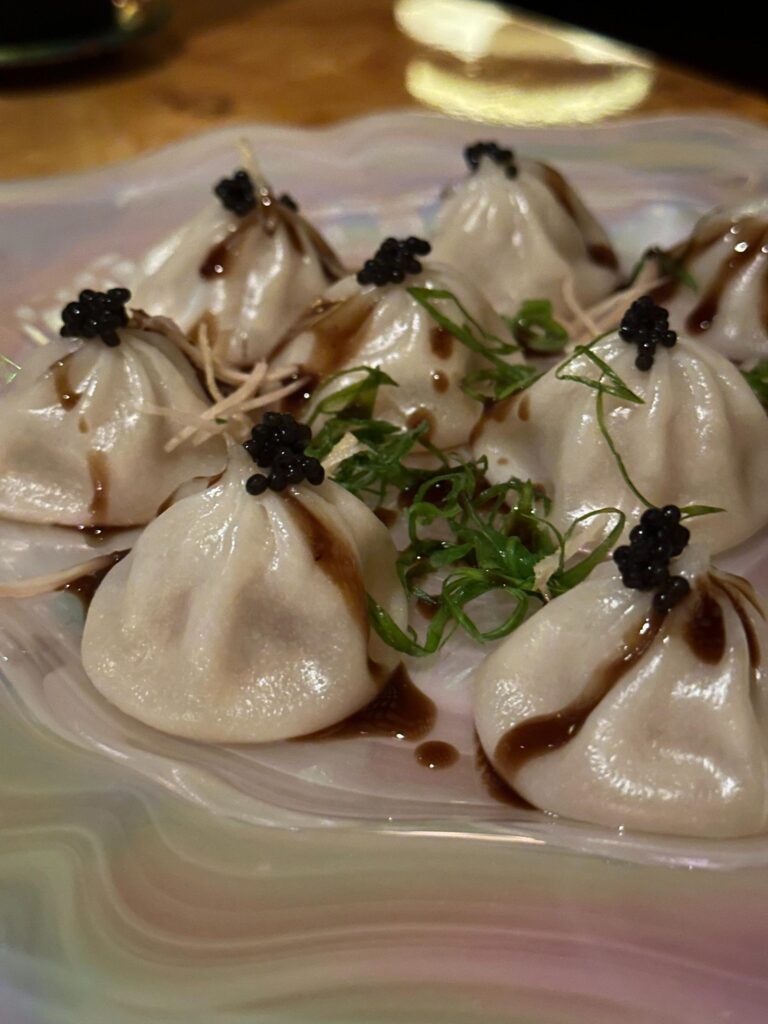 A cluster of delicate steamed dumplings is artfully plated, each drizzled with a dark savory sauce and crowned with a dollop of black caviar. Fresh scallions and ginger shreds are scattered among the dumplings on the wavy, iridescent platter, adding texture and color to the dish.