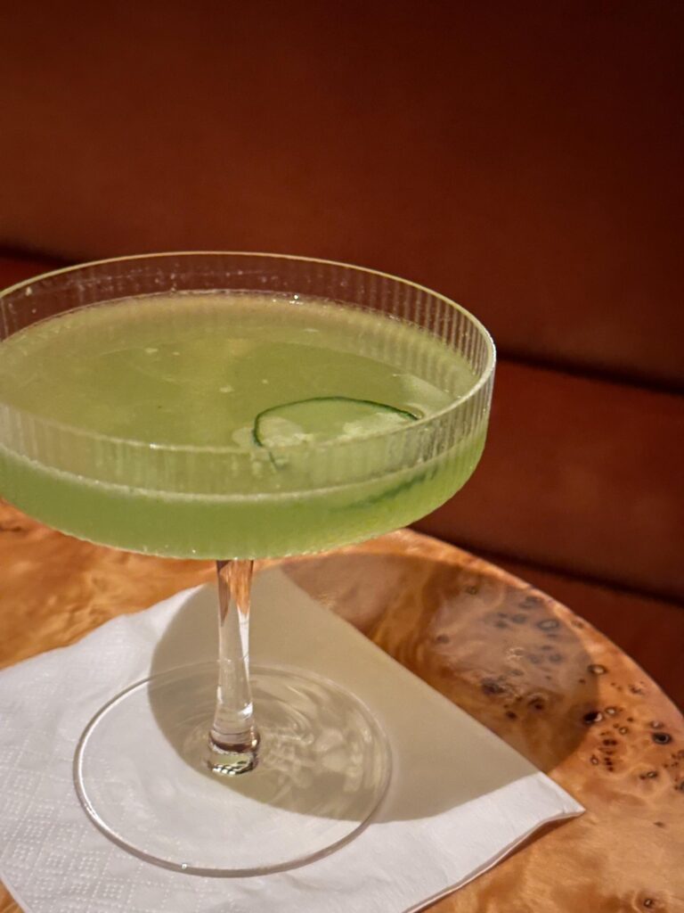 A pale green cocktail served in a textured coupe glass features a thin slice of cucumber floating on its surface. The drink rests on a white napkin atop a glossy burl wood table, set against a warm, blurred background of brown leather seating.