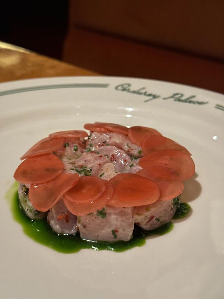 A meticulously plated circle of cubed raw fish is topped with a ring of translucent reddish slices and rests in a vibrant pool of green herb oil. The white plate features the "Corduroy Palace" name in green script along the rim, framing the appetizer against a blurred wooden table surface.