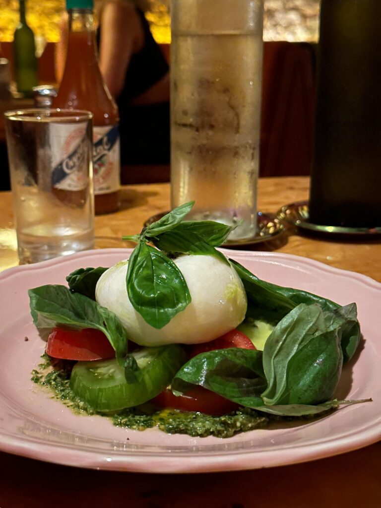 A fresh Caprese-style salad features a large ball of white cheese nestled among tomato and cucumber slices, resting on a layer of green pesto and topped with abundant basil leaves. The dish is served on a pink scalloped plate, set against a background that includes a condensation-covered glass of water and a bottle of hot sauce on a wooden table.