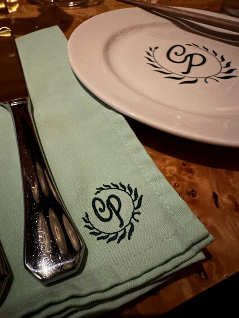 A shiny silver knife lies atop a folded mint-green napkin that features an embroidered "CP" monogram inside a laurel wreath. Next to the napkin, a white plate displaying the same dark green logo holds a pair of wooden chopsticks against the backdrop of a polished burl wood table.