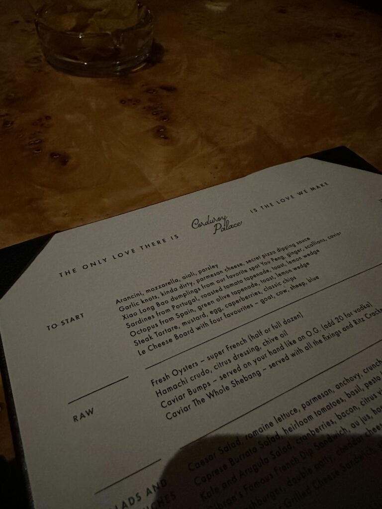 A dimly lit menu from "Corduroy Palace" rests on a glossy burl wood table, displaying sections for starters and raw dishes under the quote "The only love there is is the love we make." The visible text lists an eclectic mix of food options ranging from garlic knots to caviar bumps, with a small glass bowl of snacks blurring into the dark background.