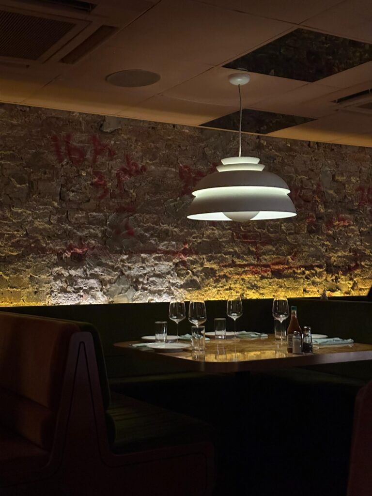 A white, tiered pendant light hangs above a table set with wine glasses and plates, which is nestled within a dark green velvet booth. The scene is framed by a rustic, distressed brick wall with red markings, illuminated by warm ambient lighting that enhances the restaurant's moody atmosphere.