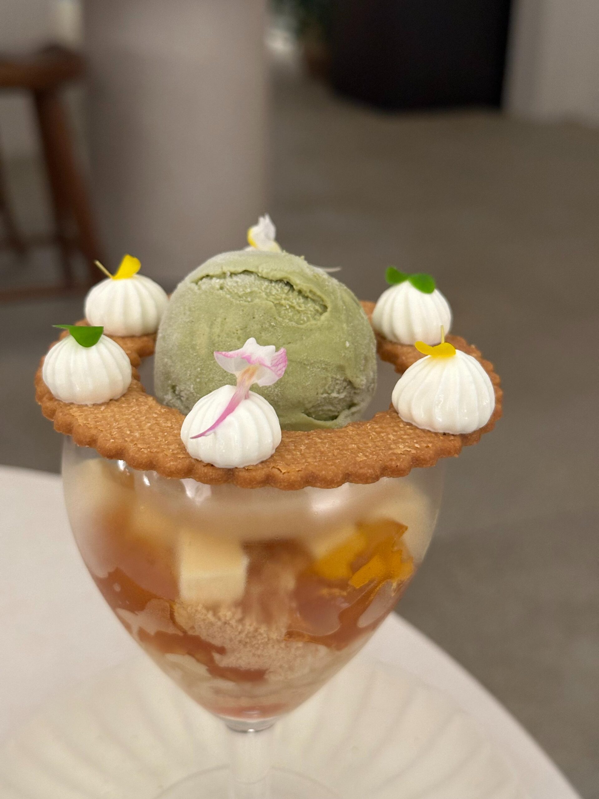 This elegant dessert features a scoop of green tea ice cream resting on a ring-shaped biscuit perched atop a glass parfait. The biscuit is decorated with delicate white meringue peaks, while the glass below is filled with layered jellies and cream.