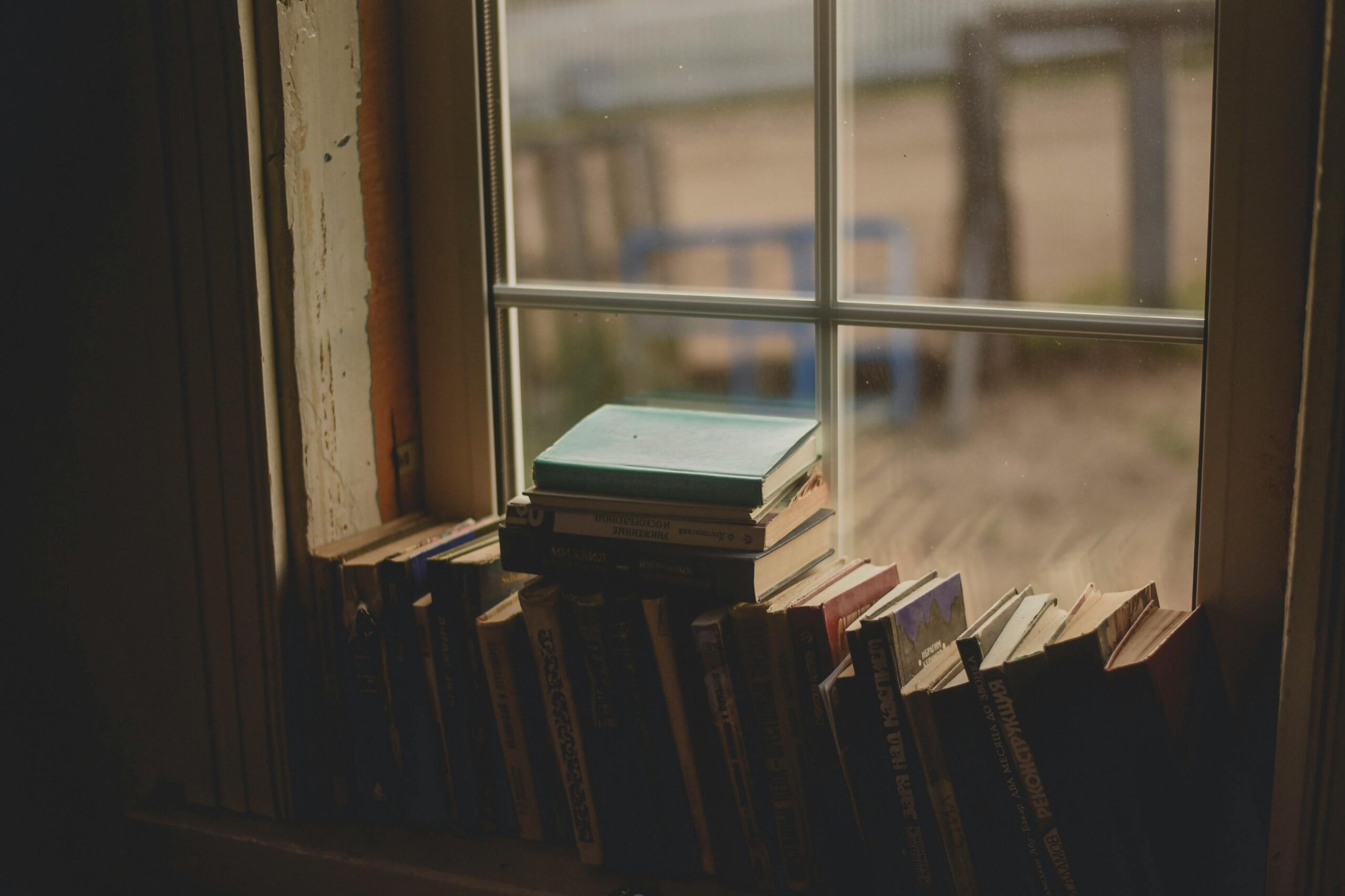 A collection of old, weathered books is lined up and stacked along a wooden windowsill, illuminated by the soft light coming through the glass panes.