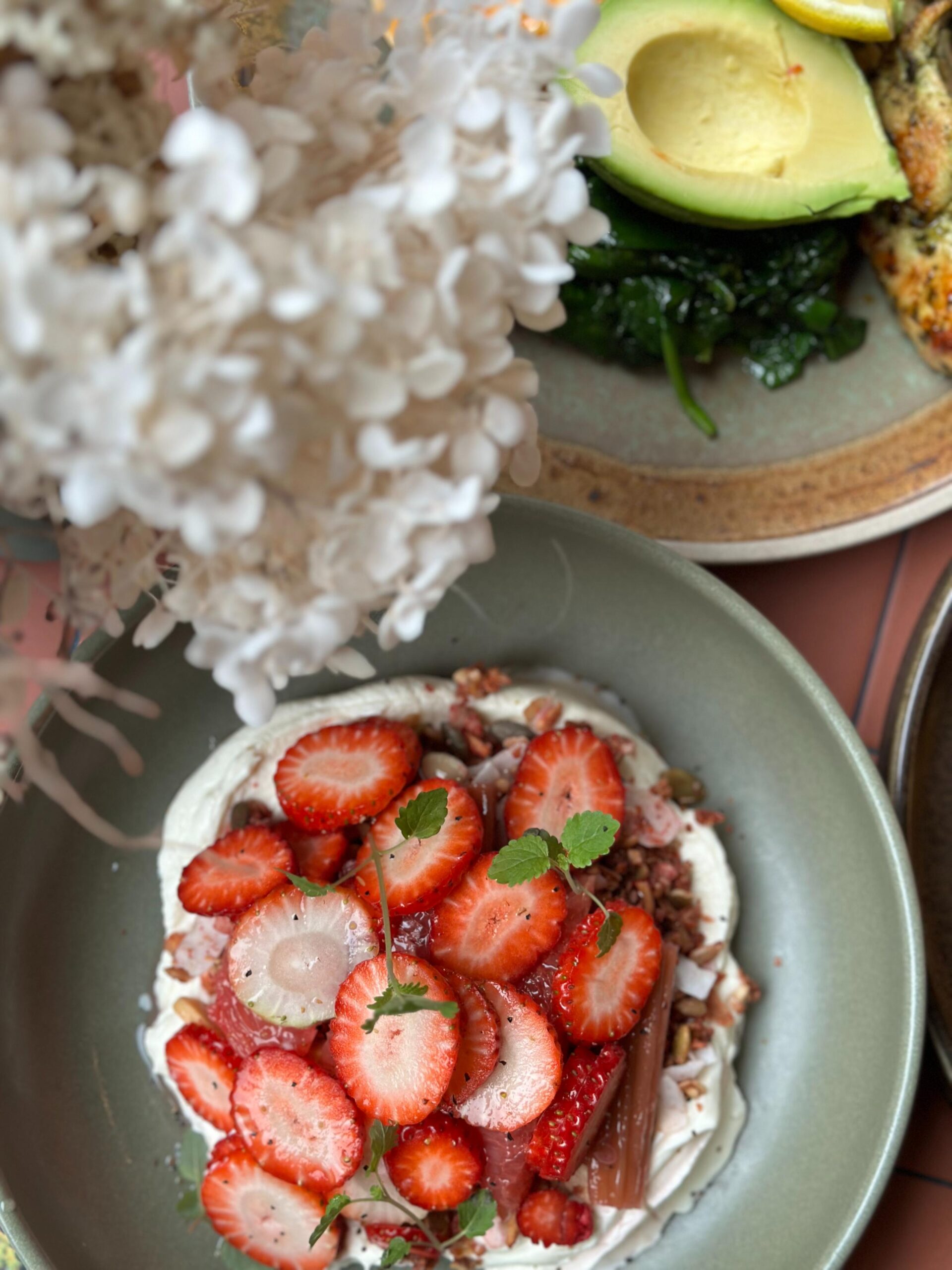 This top-down shot features a vibrant culinary spread, highlighting a bowl of creamy yogurt topped with sliced strawberries, granola, and fresh mint. In the background, a second plate offers a savory contrast with a sliced avocado, sautéed greens, and a hint of grilled protein, all framed by soft white flowers.