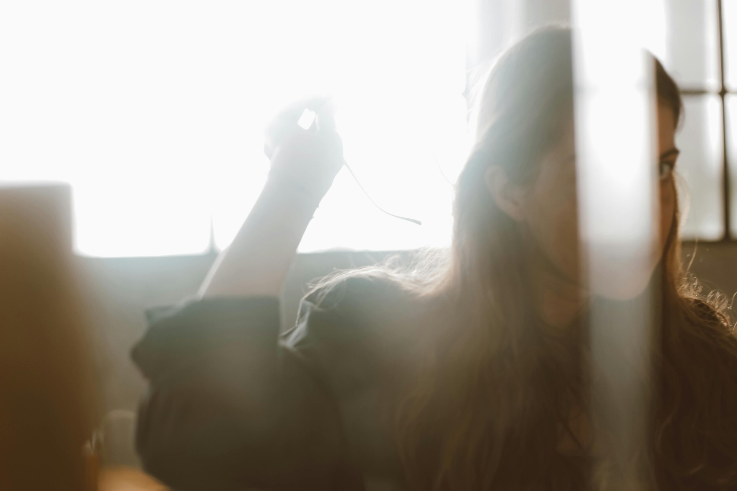 A woman with long, wavy hair is captured in a soft, ethereal light as she gazes toward the camera from behind a blurred foreground element.