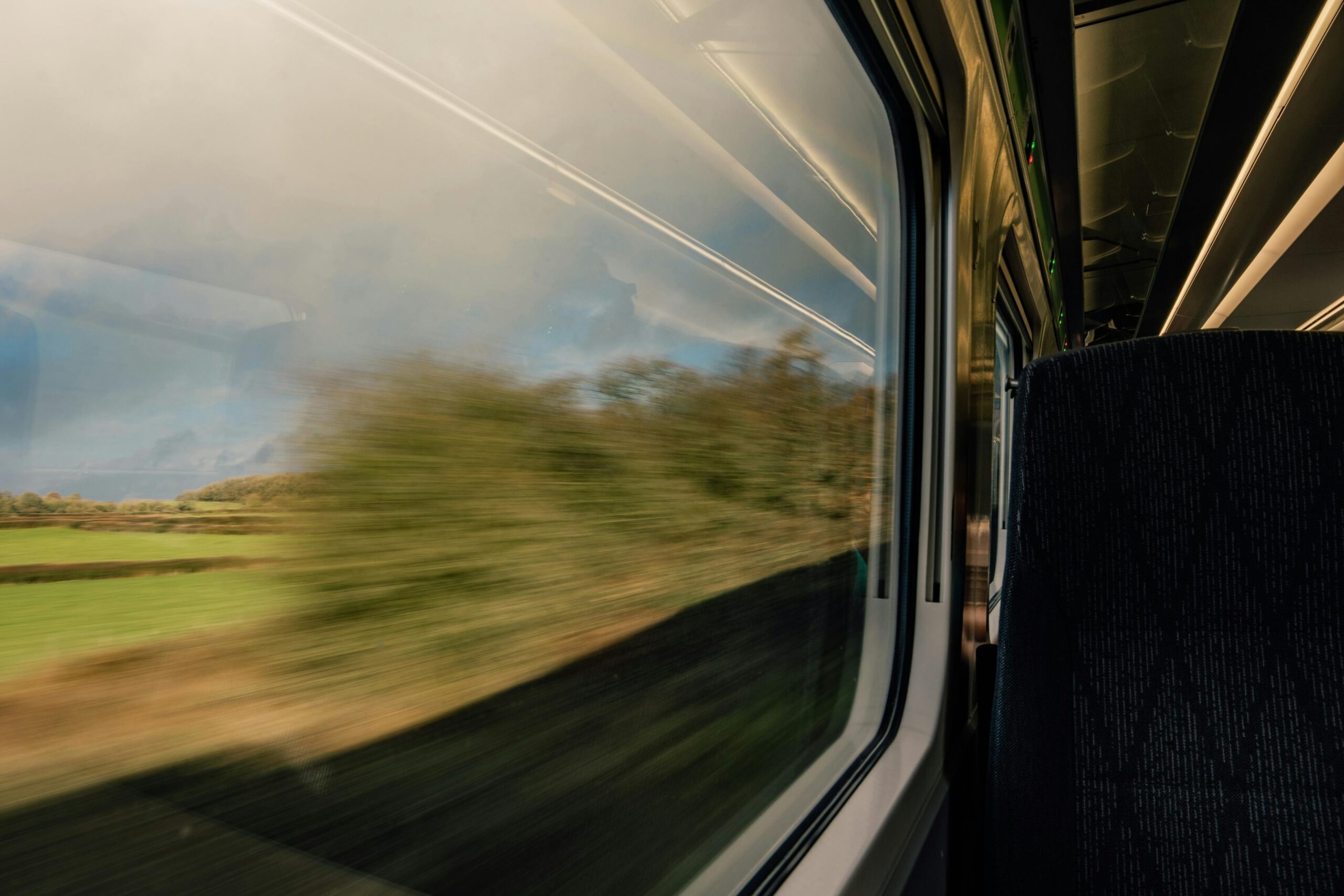 A view from inside a moving train shows a blurred green landscape rushing past a large window. The interior features a dark, patterned seat in the foreground and a warm, metallic glow reflecting off the ceiling panels.