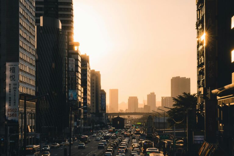 A dense flow of traffic streams through a city canyon during a hazy, golden sunset.