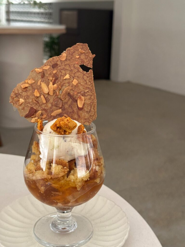 This sophisticated dessert consists of a glass goblet layered with amber-colored jelly and cake crumbles, topped with a scoop of white ice cream and crunchy honeycomb bits. A large, thin almond tuile shard is placed vertically at the back, adding height and texture to the presentation.