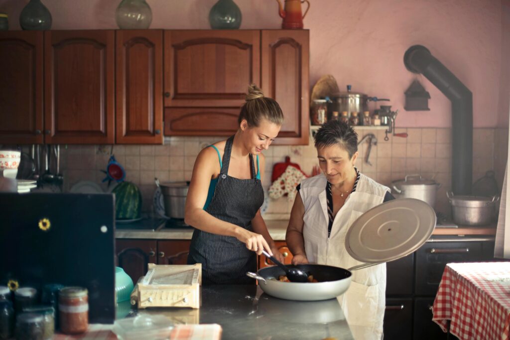 In a cozy, rustic kitchen, two women are cooking together over a pan. The younger woman is stirring the food while the older woman looks on, holding a lid in her hand.