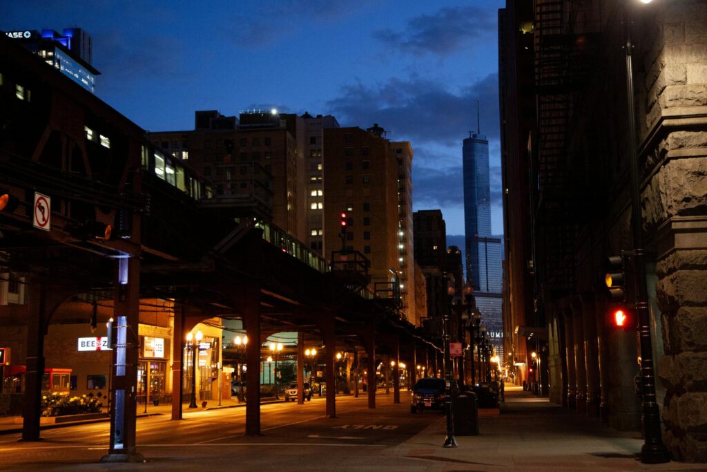 An elevated train line stretches across a dimly lit city street at dusk, illuminated by warm streetlights and glowing shop windows.