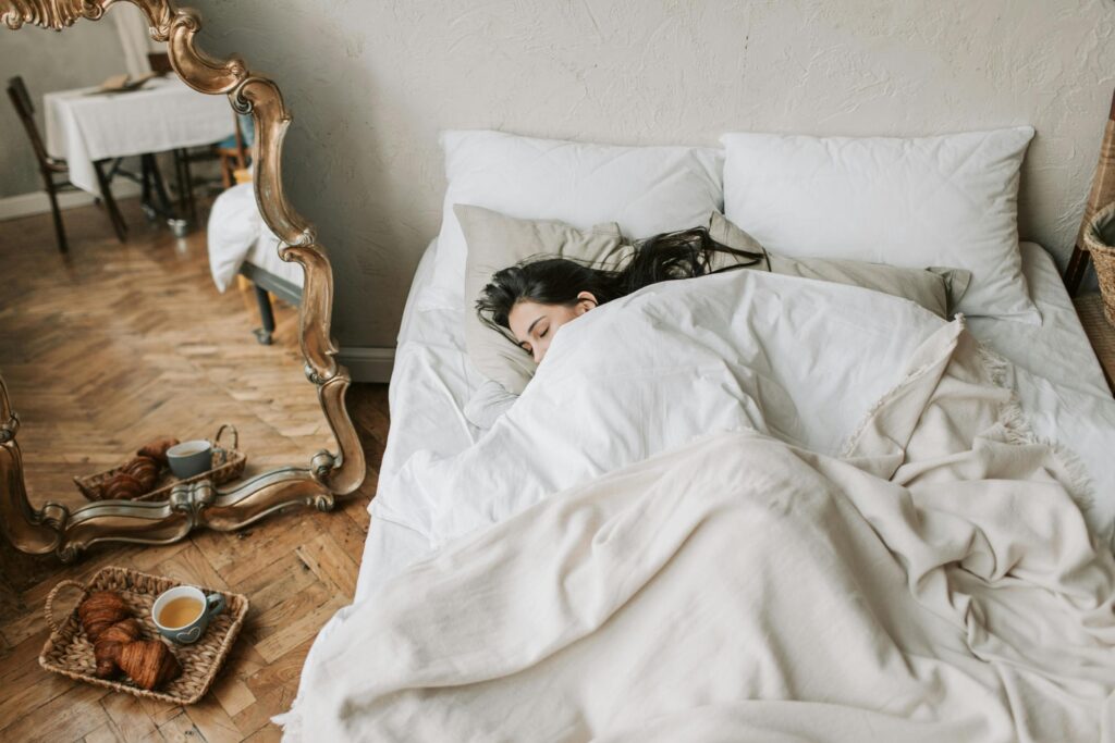 A woman sleeps soundly under white linens in a bright room, with a breakfast tray of croissants and tea resting on the hardwood floor nearby.