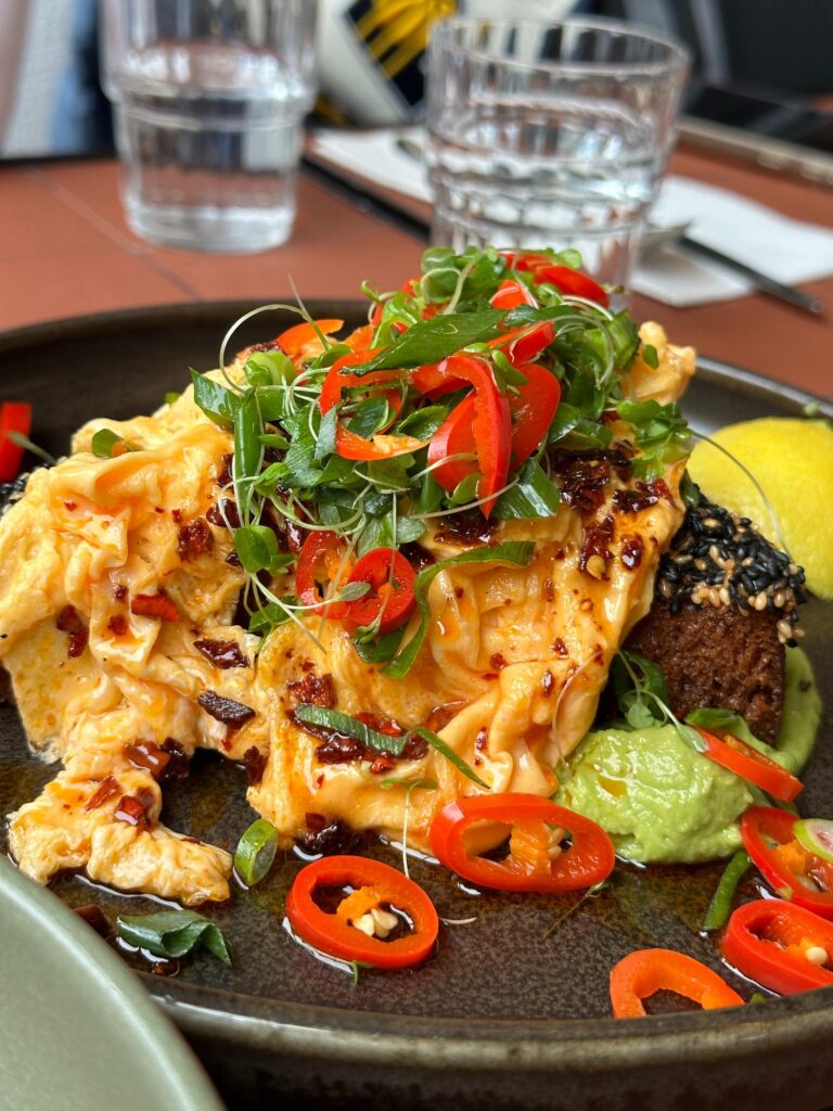 This dish features a mountain of soft-scrambled eggs draped over toast and topped with a vibrant garnish of fresh chili slices, scallions, and microgreens. The plate is finished with a side of creamy avocado mash, a sprinkle of black sesame seeds, and a wedge of lemon for a bright, flavorful meal.