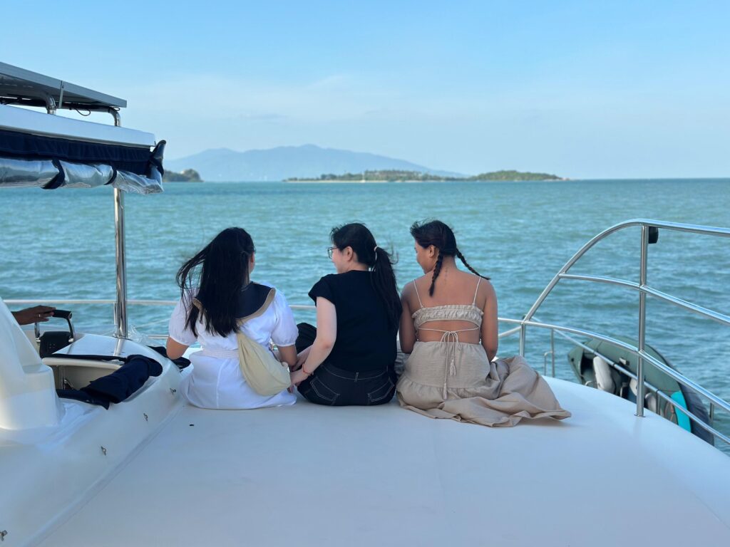 Three women sit side-by-side sitting on a yacht deck overlooking tropical islands.