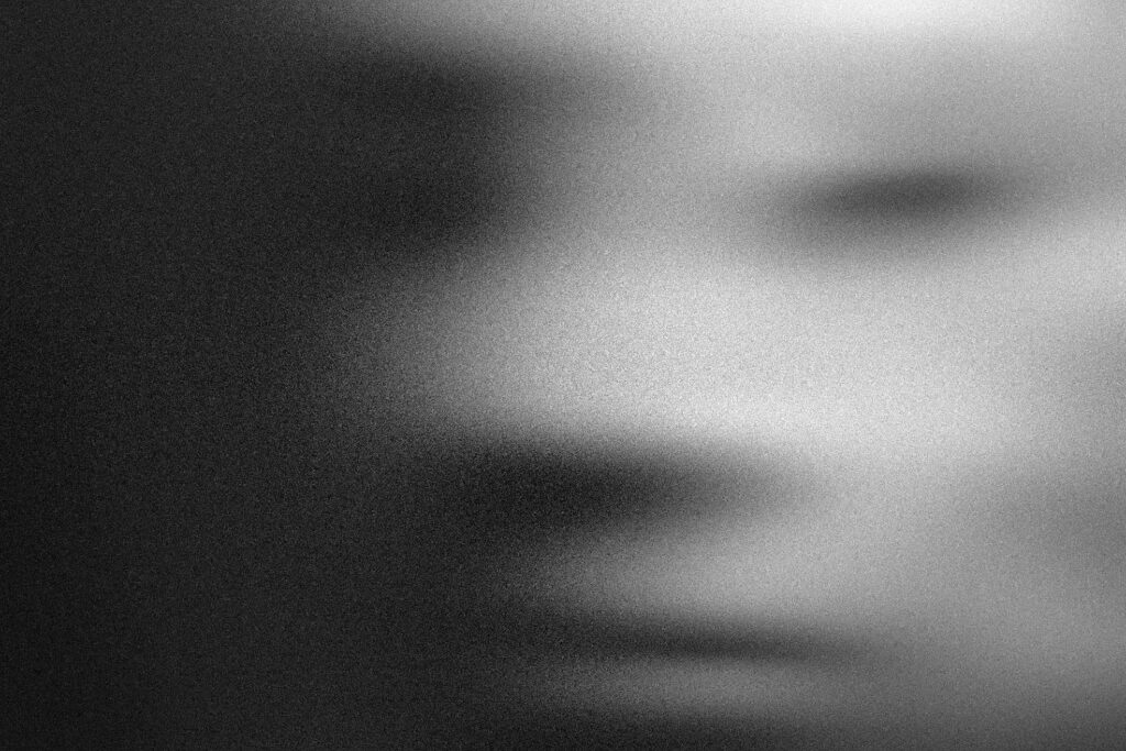 This black-and-white image features a heavily obscured and grainy close-up of a face, rendered with a soft-focus effect that blurs distinctive features.