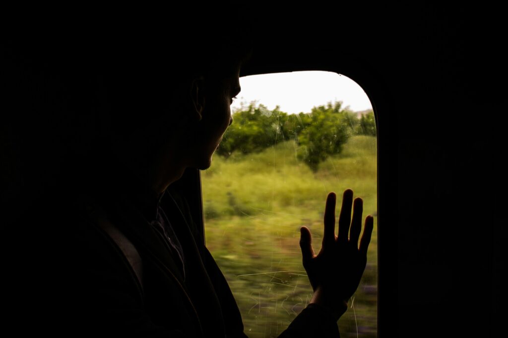 A person's silhouette is captured in a dimly lit space as they gaze out of a train window at a passing green landscape. Their hand rests gently against the glass, highlighting a quiet moment of contemplation during the journey.