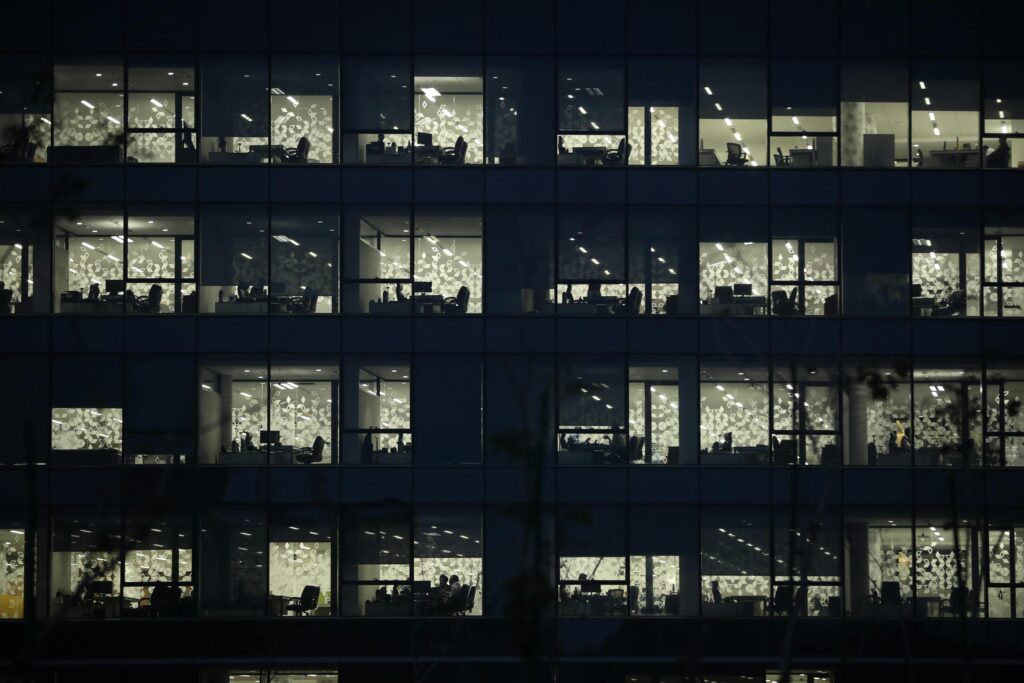 Illuminated windows reveal a grid of quiet office cubicles inside a modern building at night.
