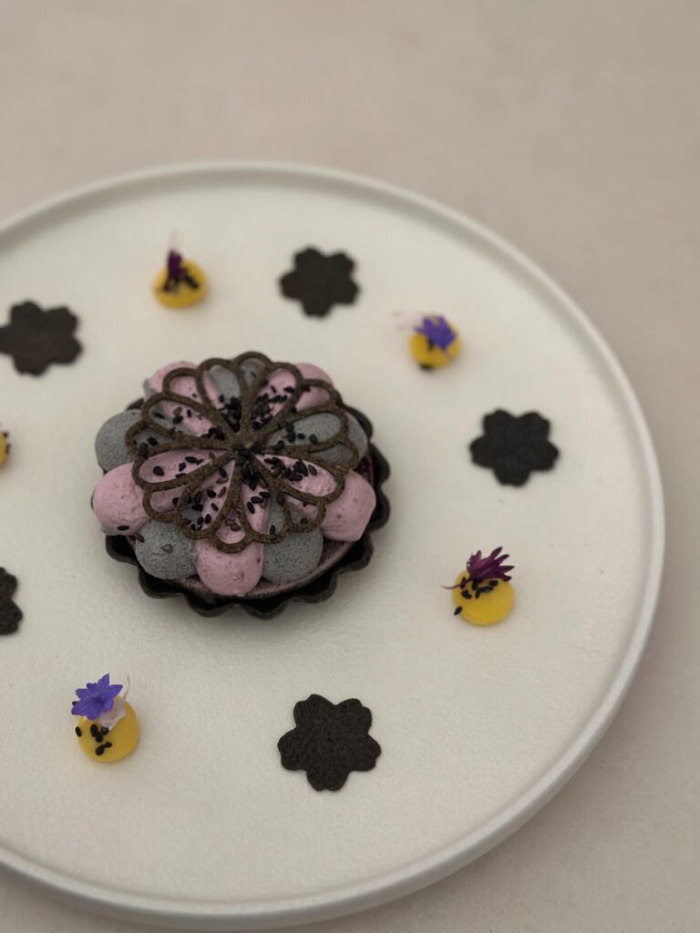 This plated dessert features a dark tart shell filled with alternating dollops of grey and pink cream, topped with an intricate chocolate filigree and black sesame seeds. The surrounding white plate is artfully decorated with yellow citrus accents, tiny purple flowers, and flower-shaped dark tuiles.