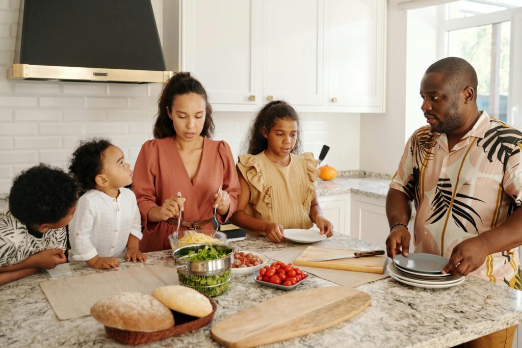 In a bright, modern kitchen with white cabinetry and marble countertops, a family of five prepares a meal together. 