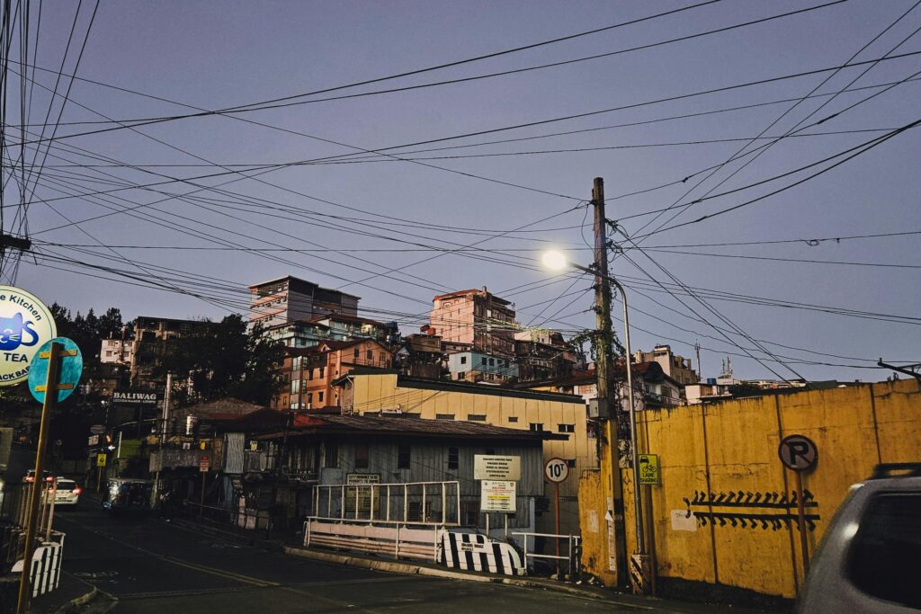 This urban street scene captures a dense cluster of buildings on a hillside under a twilight sky, framed by a complex web of overhead power lines.