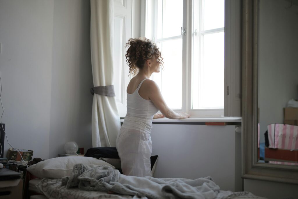 A woman with curly hair, dressed in white loungewear, stands by a bright window and gazes outside in a sunlit room.