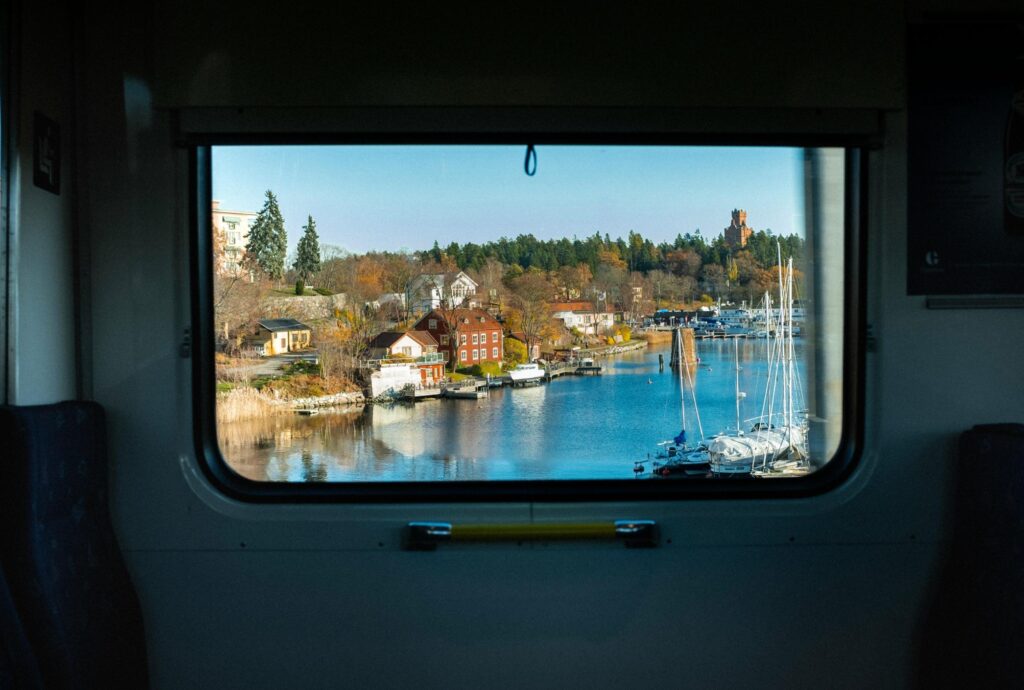 The image captures a picturesque waterfront view framed by the dark interior window of a train or bus. Outside, a cluster of colorful red and white houses sits along a blue waterway filled with sailboats and autumn trees.