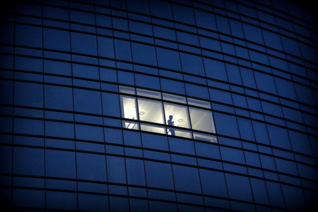 A lone silhouette stands in a brightly lit office window amidst the dark, repetitive glass facade of a skyscraper at night.