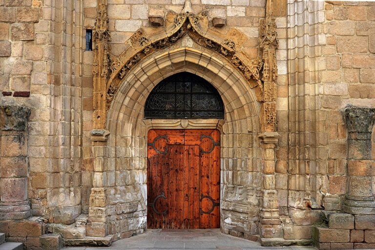 An ornate, arched stone doorway features a rustic wooden door with decorative iron hinges and a leaded glass transom above it.