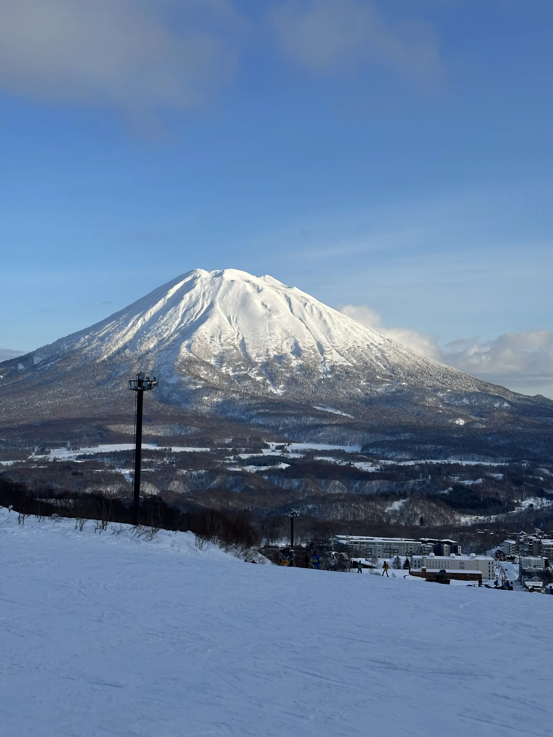 A snow-covered, symmetrical volcano towers over a winter landscape featuring a ski slope and a small village in the valley. The scene is set under a clear blue sky, highlighting the rugged textures of the mountain’s peak and the surrounding evergreen forests.
