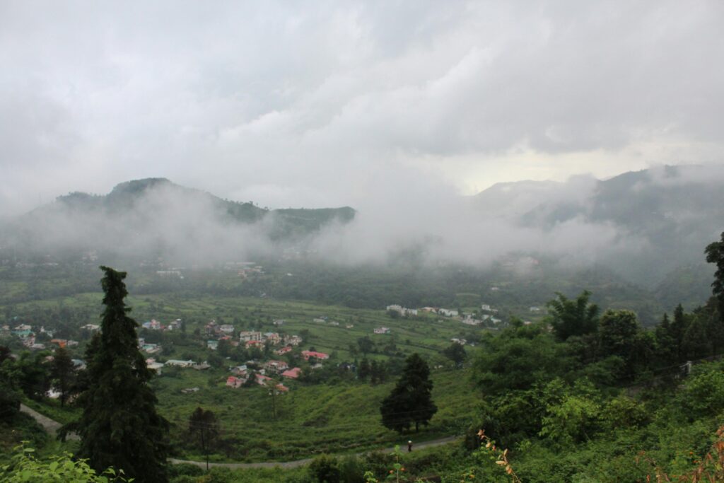 Thick, low-hanging clouds and mist drift across a lush, green mountain valley dotted with small houses. The scene is captured from an elevated perspective, overlooking a winding road and dense clusters.