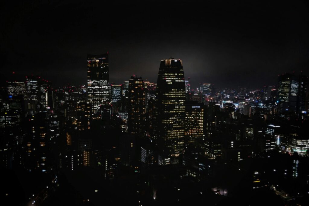 This night photograph captures a sprawling city skyline illuminated by countless glowing windows and red aviation lights against a dark, moody sky. In the center, a prominent, rounded skyscraper stands tall as the focal point among a dense sea of shorter urban buildings.