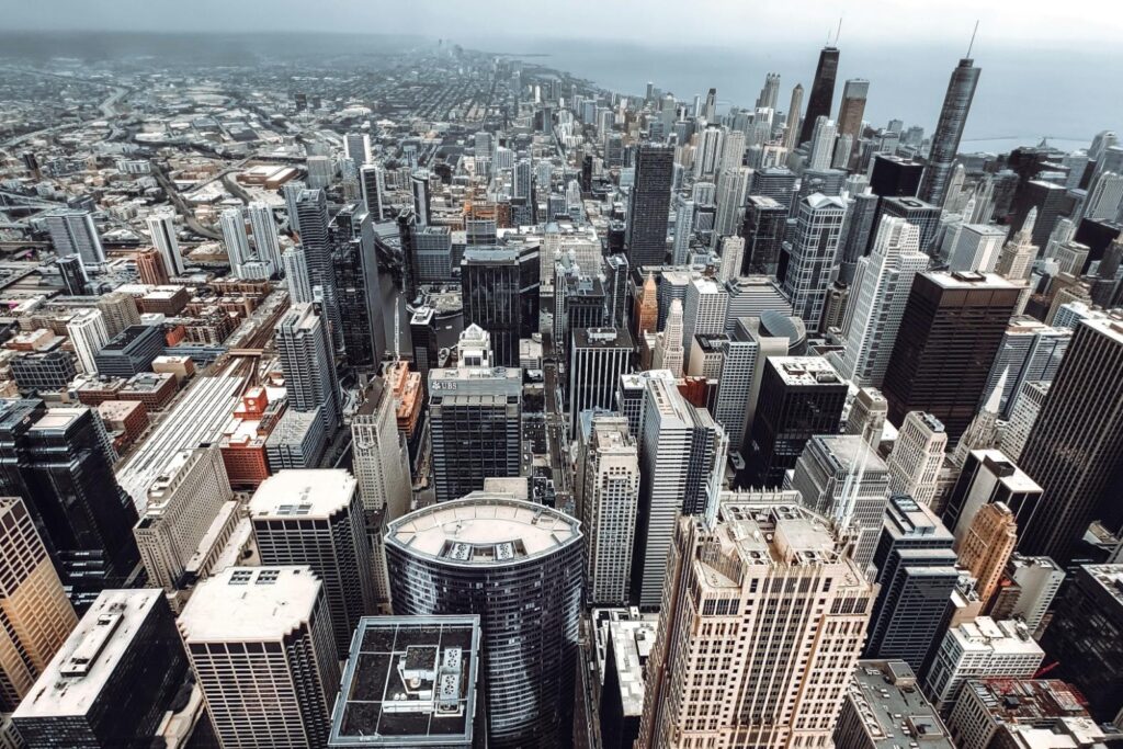 This high-angle aerial photograph captures the dense, sprawling skyline of Chicago on an overcast day. The image features a vast array of architectural styles, with prominent skyscrapers.