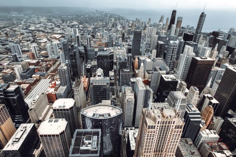 This high-angle aerial photograph captures the dense, sprawling skyline of Chicago on an overcast day. The image features a vast array of architectural styles, with prominent skyscrapers.