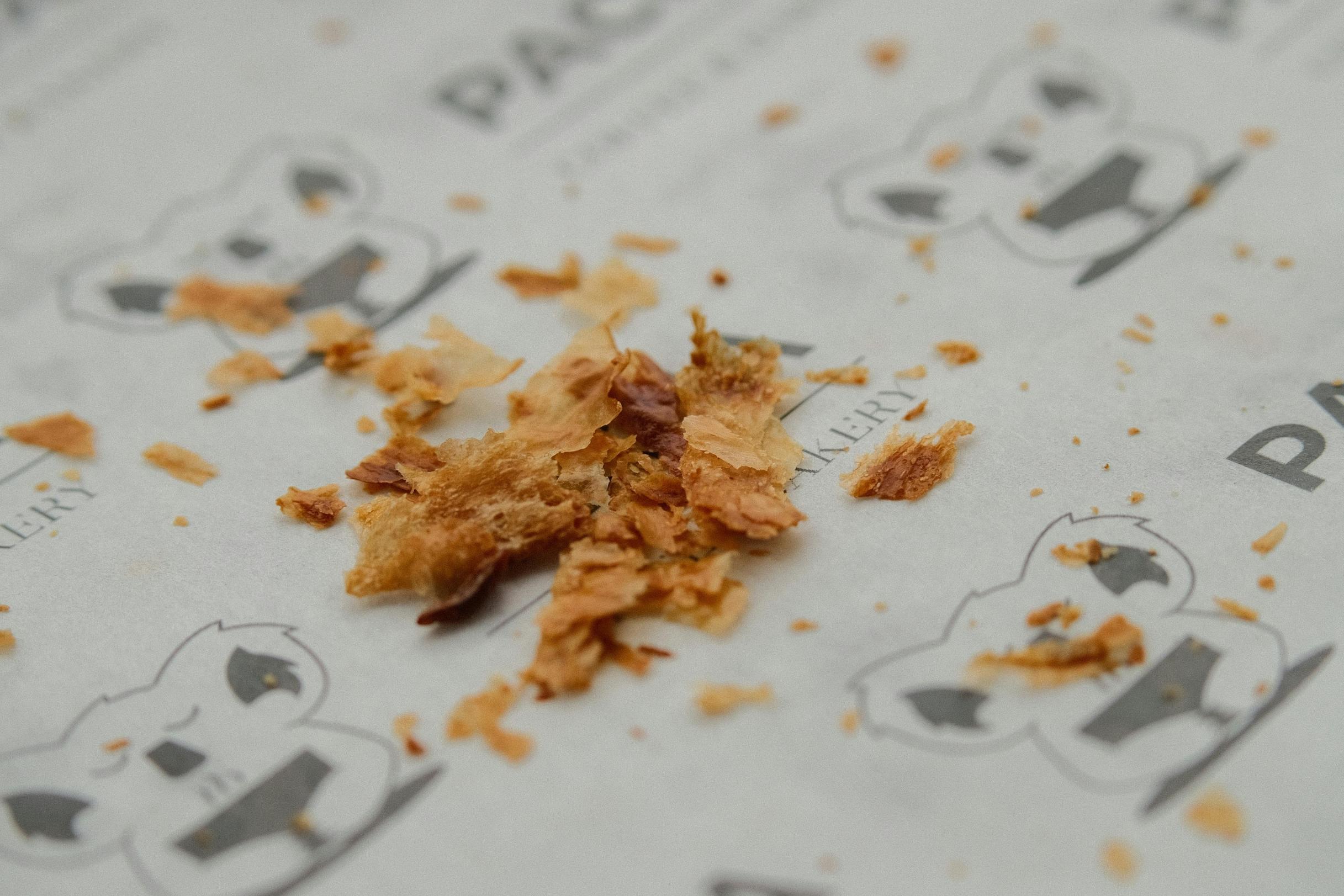 Golden, flaky crumbs from a pastry are scattered across a piece of white parchment paper. The paper features a repetitive grey logo of a koala head accompanied by the word "BAKERY."