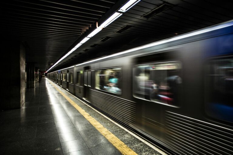 A subway train speeds through a dark underground station, captured as a metallic blur against the platform's yellow safety line. Overhead fluorescent lights create long, glowing streaks.