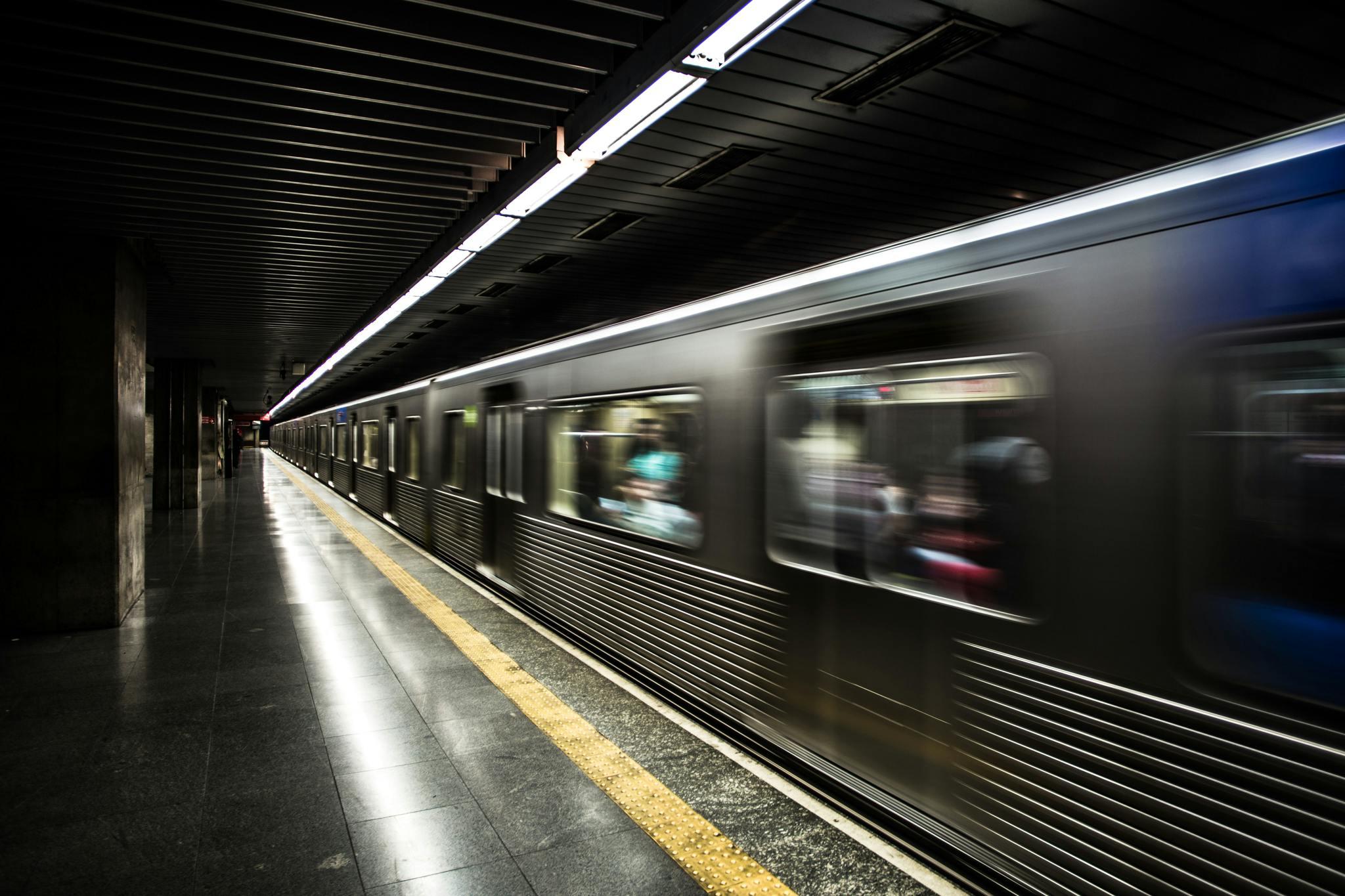 A subway train speeds through a dark underground station, captured as a metallic blur against the platform's yellow safety line. Overhead fluorescent lights create long, glowing streaks.