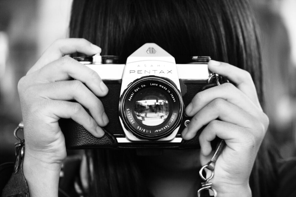 A person holds a vintage Asahi Pentax film camera up to their face, obscuring their features while focusing through the lens. The black-and-white photograph emphasizes the metallic textures of the camera and the detailed markings of the Super-Takumar lens.