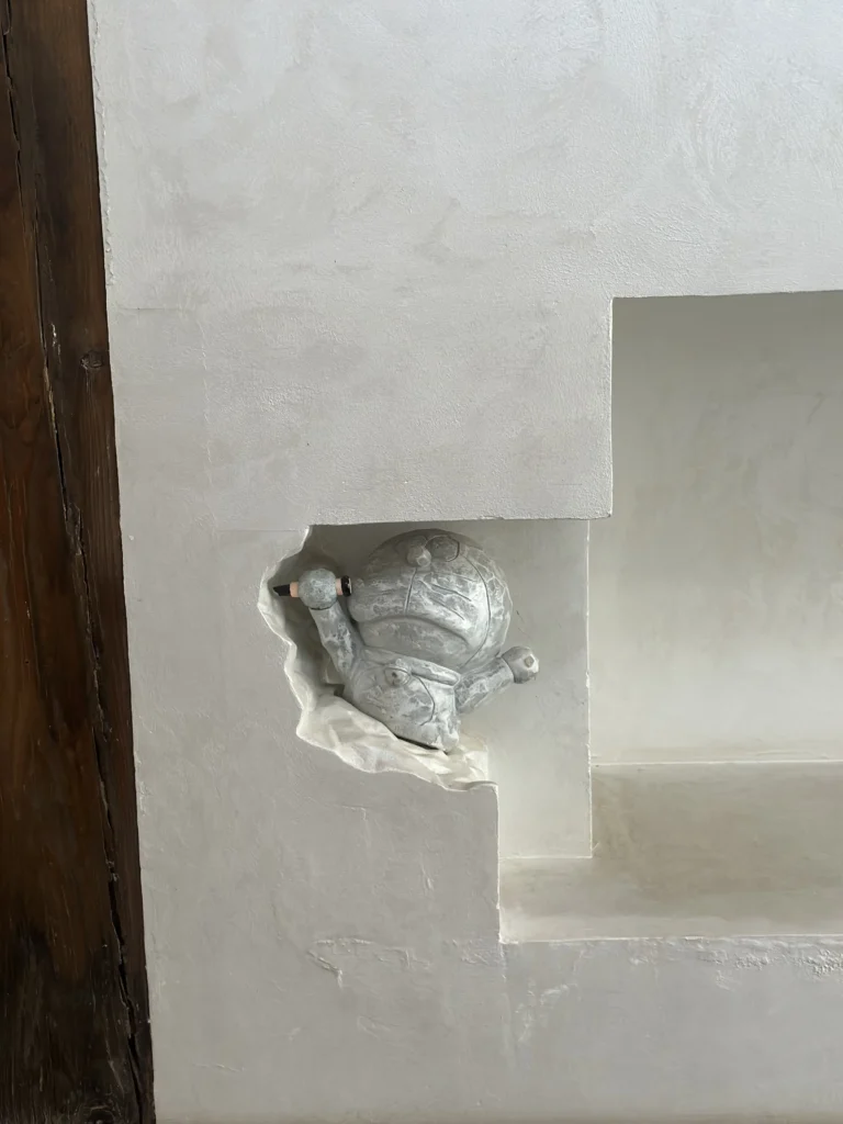 A weathered, stone-like statue of the character Doraemon appears to be breaking through a jagged hole in a white, textured wall. The figure is posed playfully within a minimalist architectural niche, creating a whimsical contrast between the modern interior and the classic pop-culture icon.