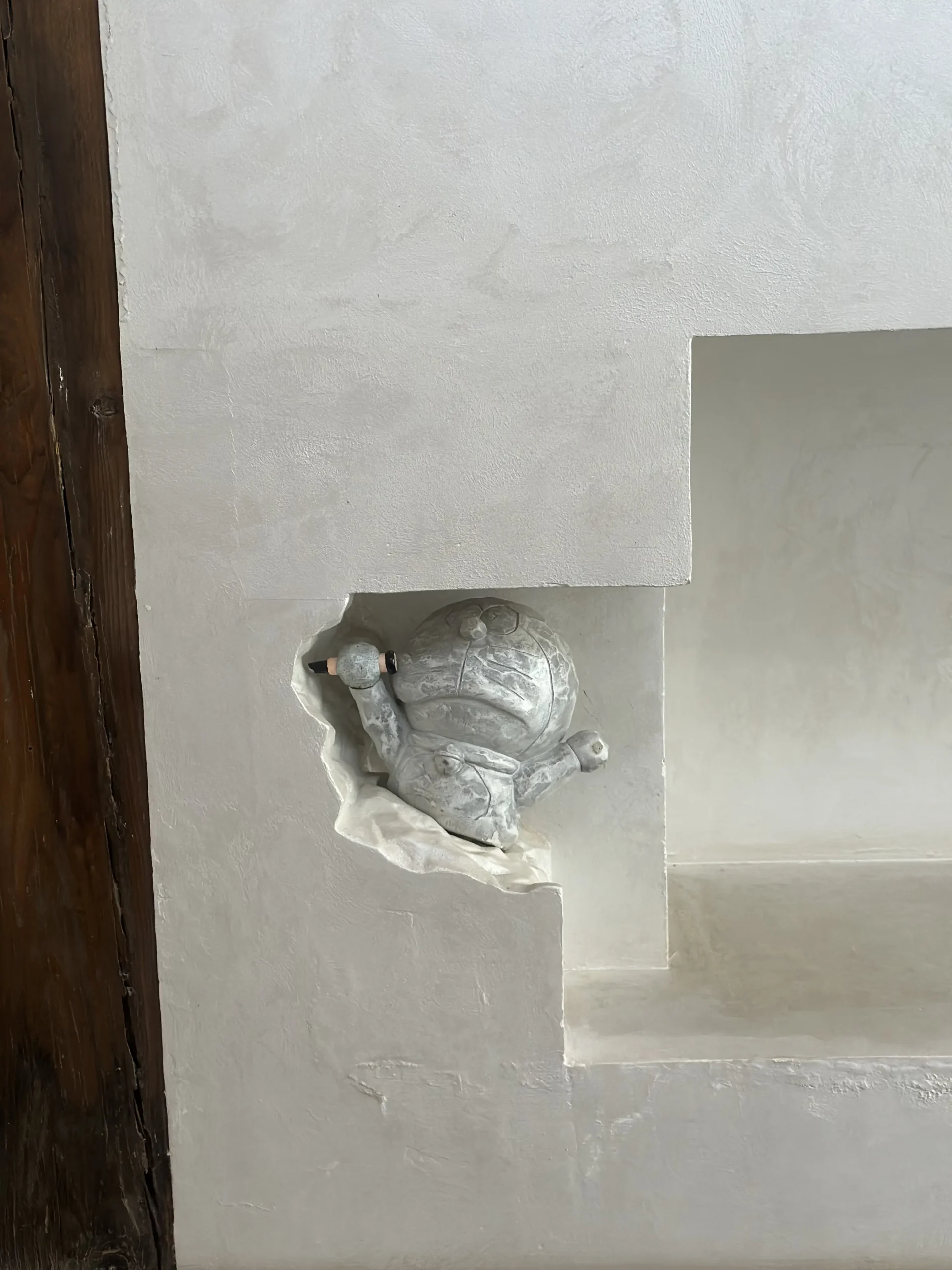 A weathered, stone-like statue of the character Doraemon appears to be breaking through a jagged hole in a white, textured wall. The figure is posed playfully within a minimalist architectural niche, creating a whimsical contrast between the modern interior and the classic pop-culture icon.